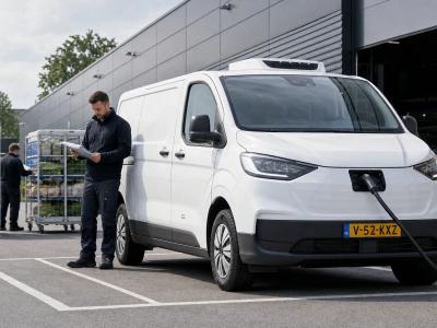 Een brede buitenopname van een elektrische bestelwagen in stedelijke distributie, geparkeerd bij een laadpunt aan de rand van een magazijnzone. Op de achtergrond staan een paar koelkarren met verse producten en een tweede medewerker die een routecheck doet op een papieren planner naast de wagen. De laadruimte is gesloten, zodat de focus ligt op de combinatie van elektrisch rijden, gekoeld transport en dagelijkse inzetbaarheid voor korte leveringen.