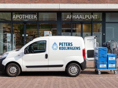 Een compacte koelbus staat bij een apotheek en afhaalpunt met kratten voor gekoelde bezorging in de buurt.