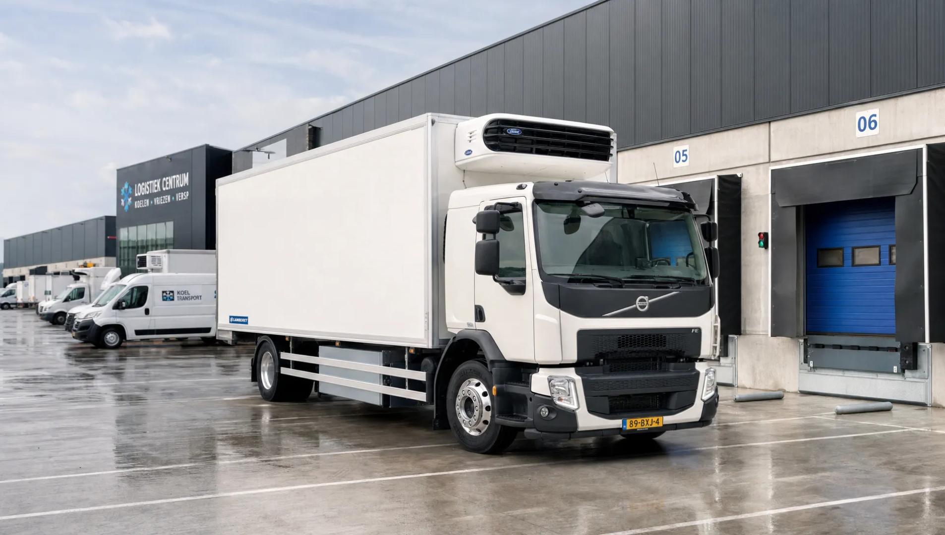 Een transporter koelwagen staat geparkeerd op een logistiek terrein bij laadperrons en andere bestelwagens.
