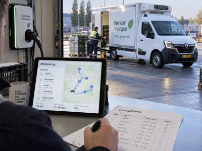 Een planner bereidt een route voor naast een laadstekker, terwijl een elektrische koelwagen klaarstaat bij de distributiehal.
